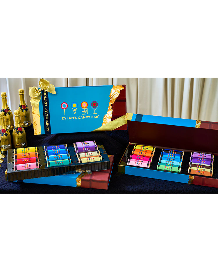 3 Anniversary Edition XL Chocolate Bar Gift Sets on a table. One box is closed with chocolate bars on top. One box is open and displaying chocolate bars. One box is closed and propped up. Candy champagne bottles are in the back. 